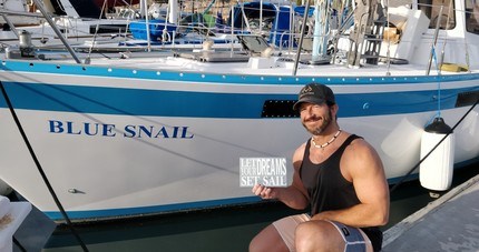 Keith in front of his sailboat 'Blue Snail', with a sign that says 'Let Your Dreams Set Sail'